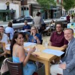 People dining at tables in a curbside outdoor plaza beside a city street