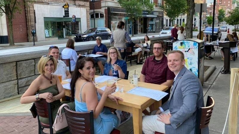 People dining at tables in a curbside outdoor plaza beside a city street